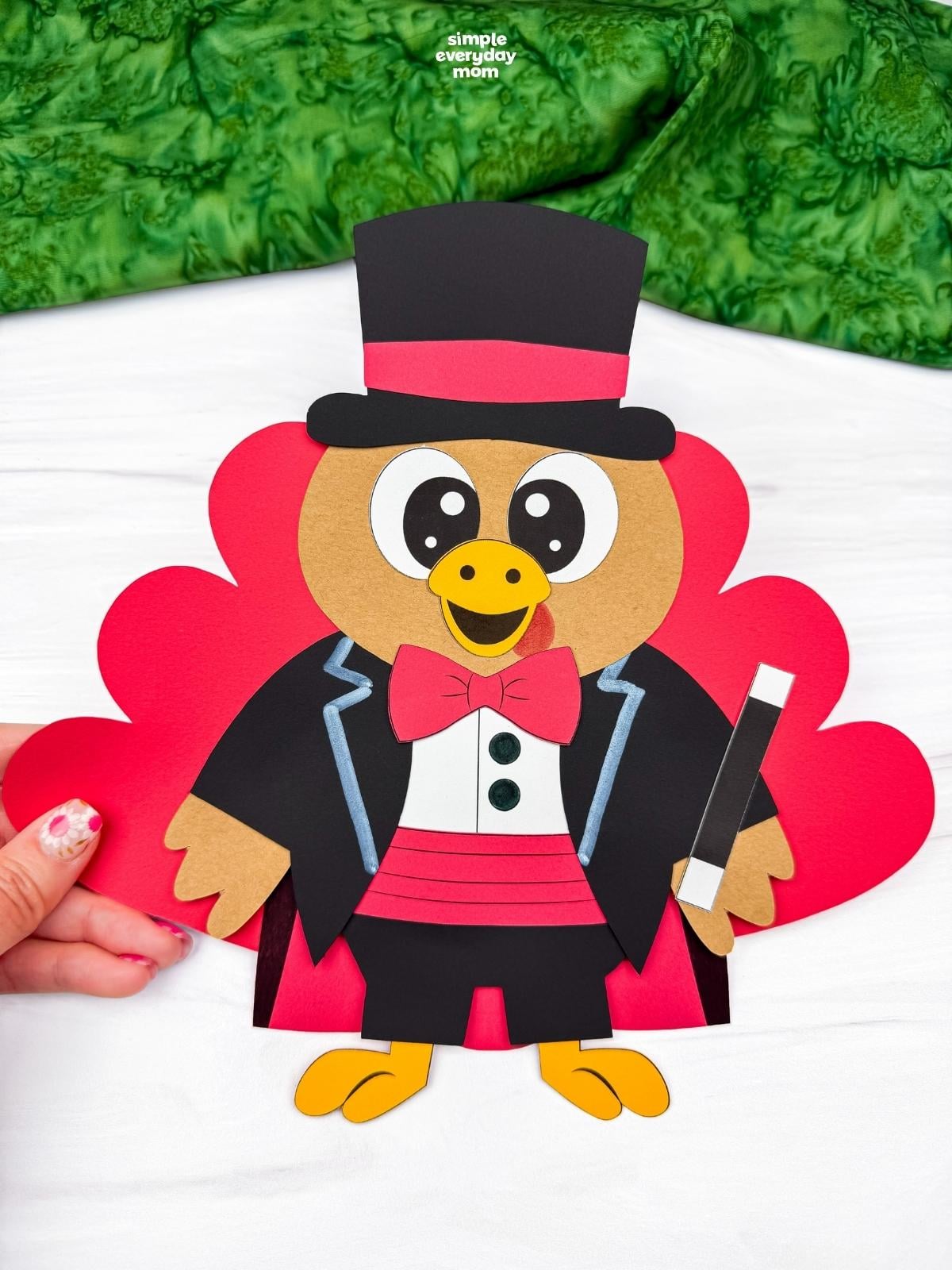 Hands holding a completed Magician Turkey Disguise Craft featuring red tail feathers, a black top hat, red bow tie, black jacket, and magic wand.
