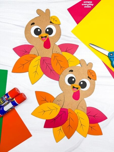 A completed craft featuring two cute fall paper turkeys nestled in colorful autumn leaf piles, surrounded by construction paper, glue sticks, and scissors.