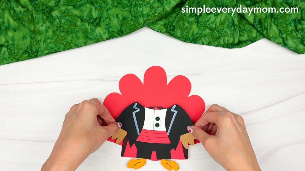 Hands positioning the turkey's body over the red tail feathers for the Magician Turkey Disguise Craft.