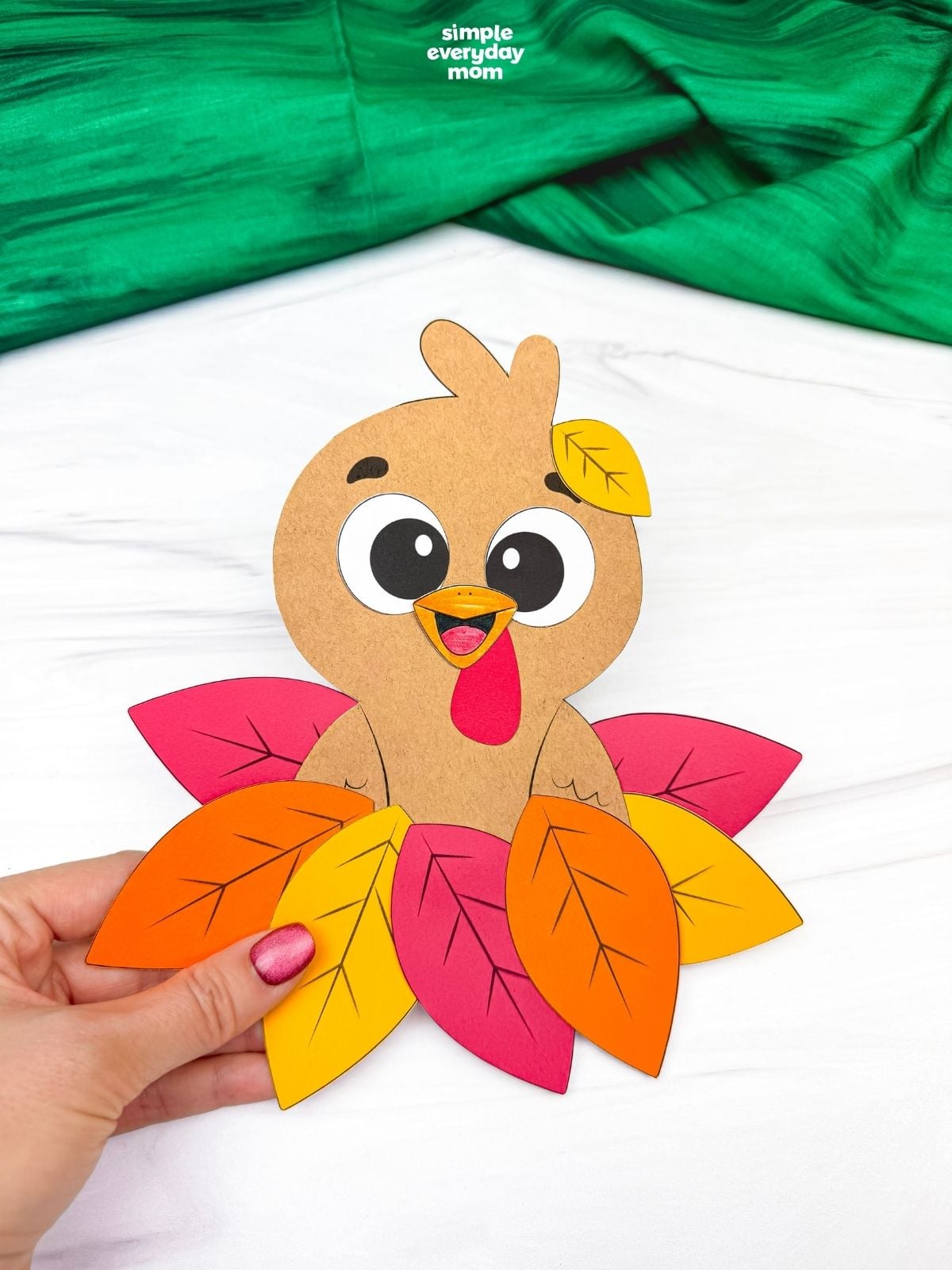 Hands holding a completed fall turkey paper craft featuring a cute brown turkey surrounded by colorful autumn leaves.