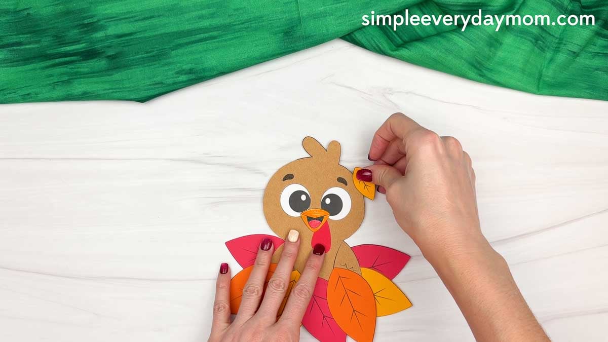 Hands attaching a small autumn leaf to the side of the turkey's head to complete the fall turkey paper craft.
