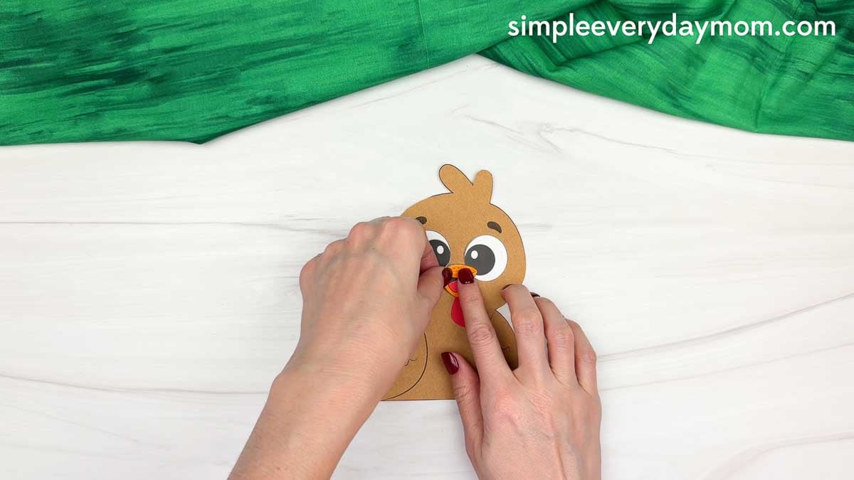 Hands gluing the orange beak onto the turkey's face above the red snood for a fall turkey paper craft.