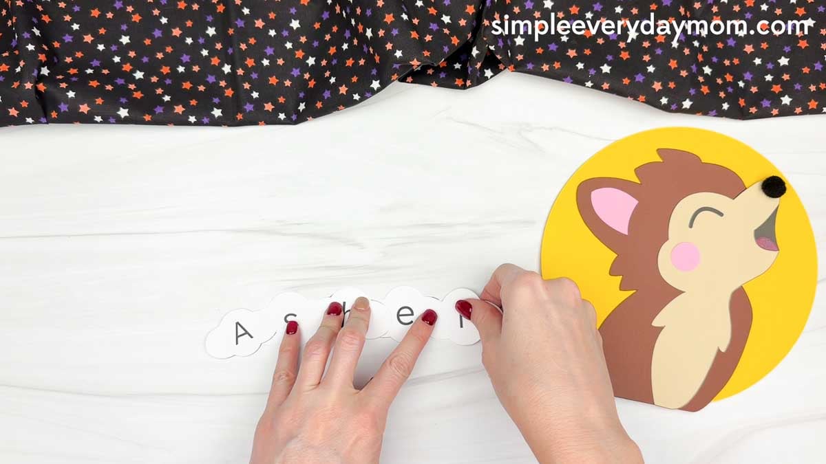 Hands arranging lettered moon pieces to spell a child's name beside the werewolf name craft for Halloween.