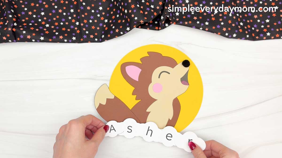 Hands positioning the lettered pieces spelling a child's name along the bottom of the werewolf name craft for Halloween.